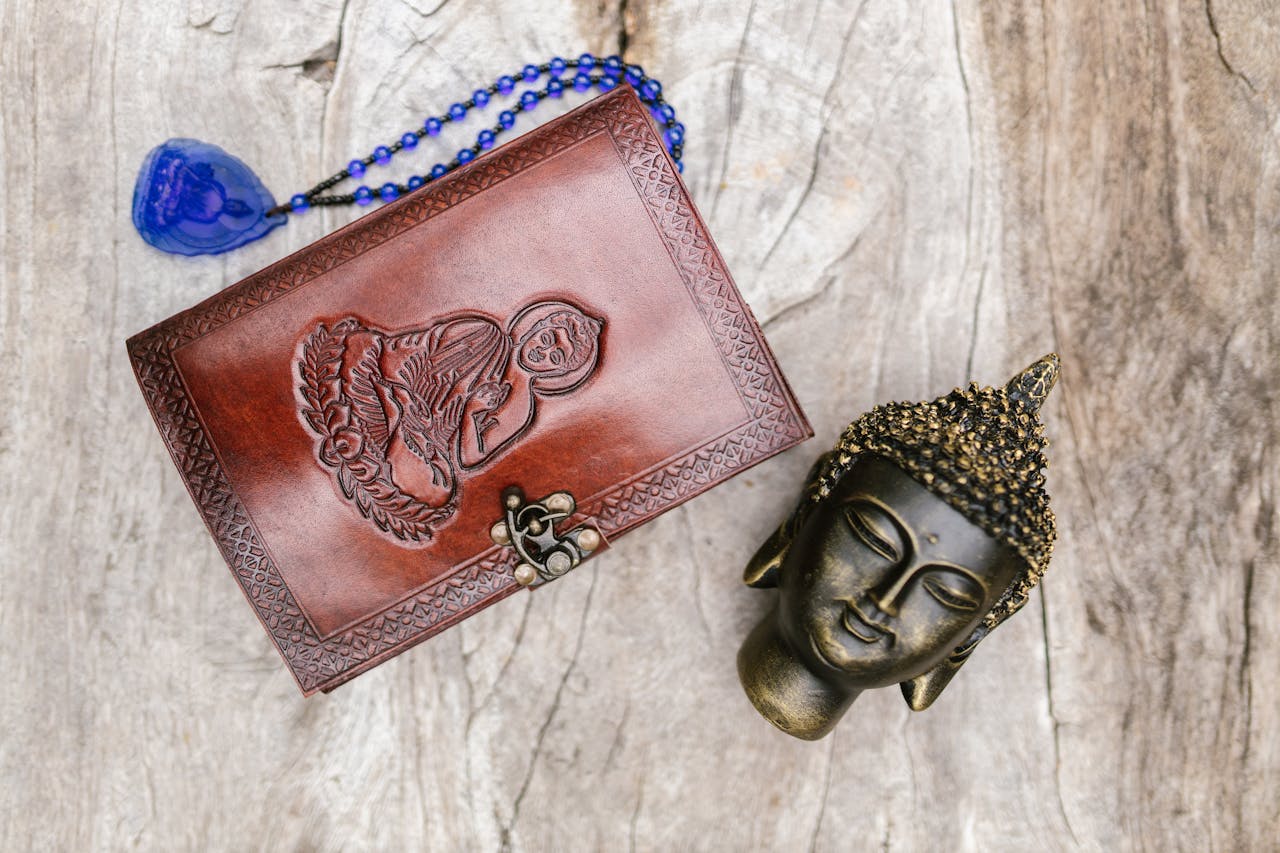 Buddha head sculpture and engraved leather book with prayer beads on rustic wooden table.